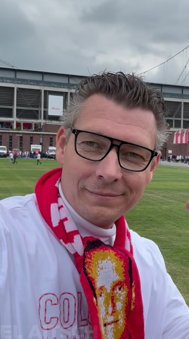 Lars Hüsemann am Aufstiegstag mit rot-weißem Schal auf dem Rasen des RheinEnergieStadions, im Hintergrund die Tribüne.
